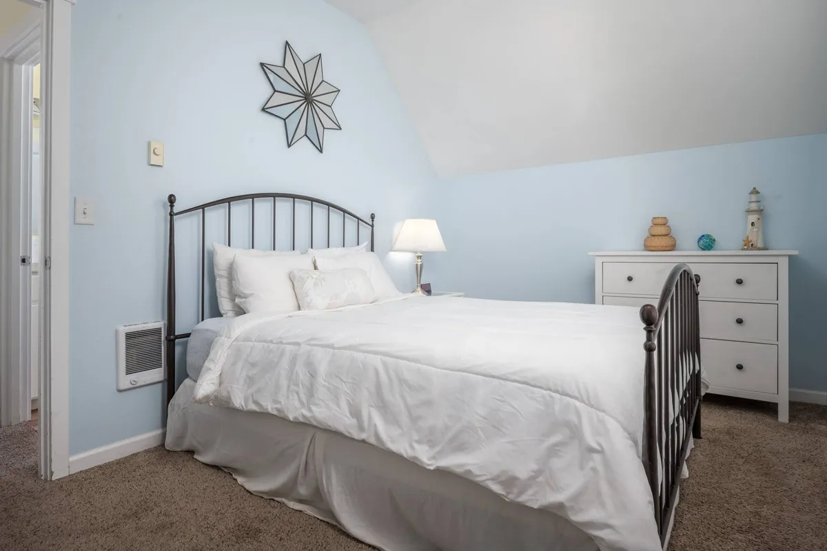 A tranquil bedroom featuring a plush bed, nightstand with a lamp, a stylish dresser, and a decorative star. Natural light pours in, enhancing the serene and quiet ambiance.