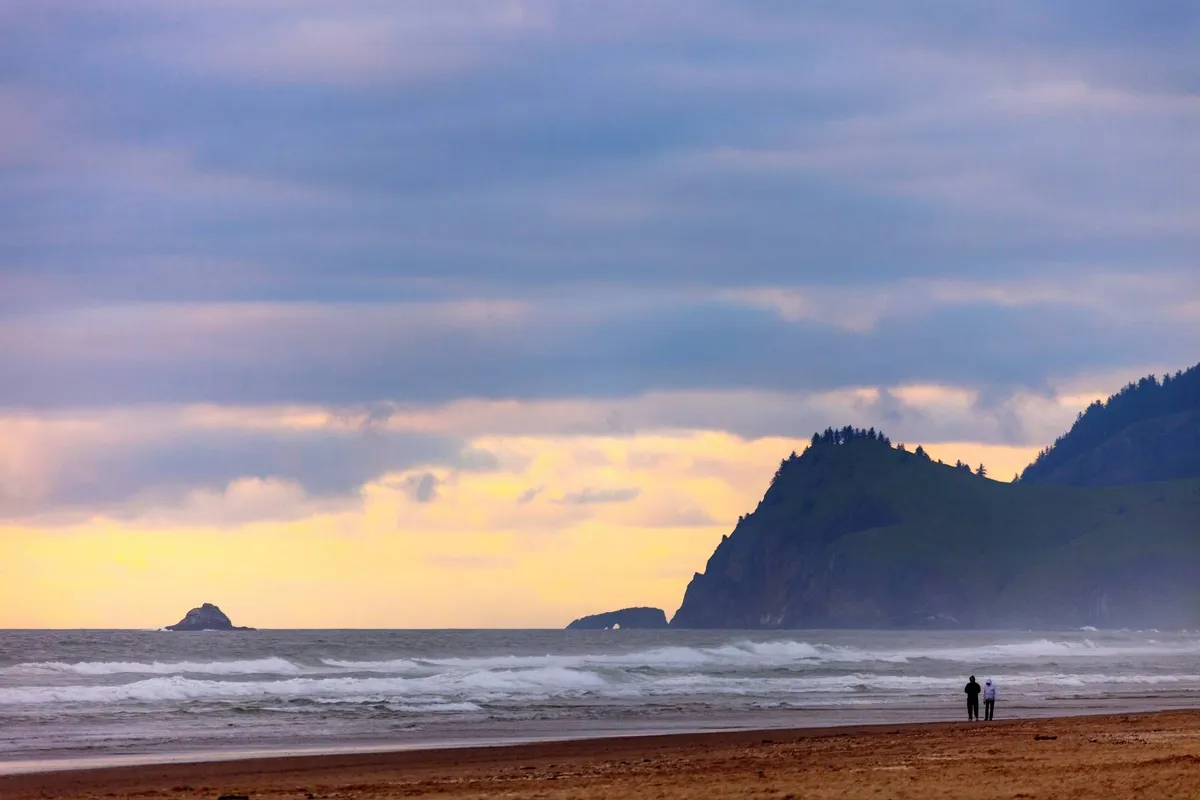 A couple walks together on a sandy beach, with gentle ocean waves, rock formations, and distant cliffs in the background. The scene is tranquil and reflective, perfect for peaceful getaways or family adventures.