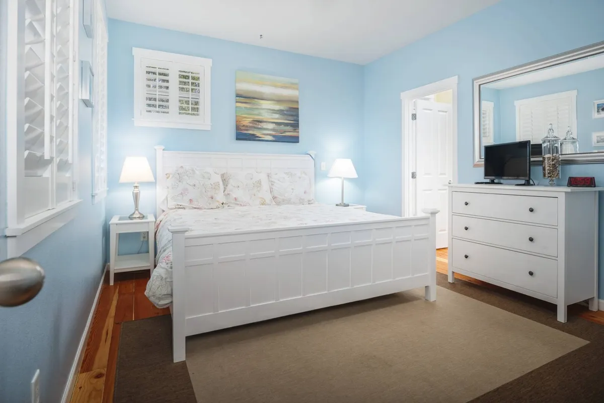A serene bedroom featuring a comfortable bed, dresser, tasteful artwork, and soft lighting from matching lamps. The soft morning light filters in, creating a peaceful atmosphere ideal for rest and relaxation.