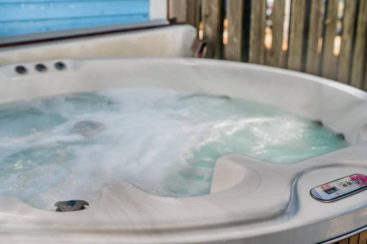 A private hot tub bubbling invitingly in a fenced backyard. The setting is tranquil, with enough privacy for a relaxing soak, and greenery at the edges suggesting a pleasant garden atmosphere. The control panel and gentle lighting imply it’s ready for starlit evenings or unwinding after a day of activity.