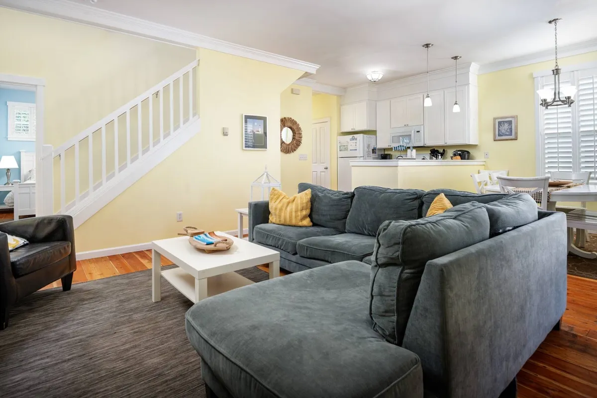 A spacious, open-concept interior featuring a large sectional sofa arranged around a wooden coffee table. The background reveals the adjoining kitchen area and a staircase, all bathed in natural light from large windows. The atmosphere is inviting and perfect for gatherings.