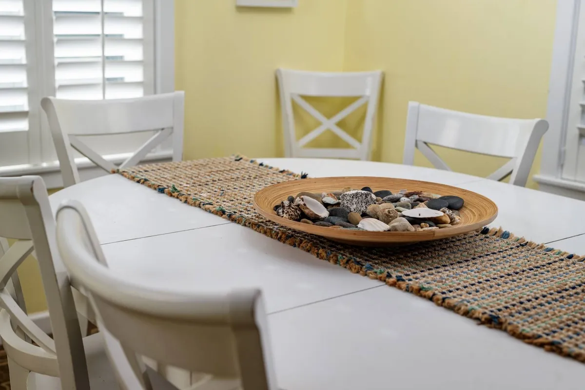A bright and welcoming dining area with a large table set for casual meals. White dining chairs surround the table, which features a decorative bowl of stones and a woven runner, suggesting space for groups to gather, share meals, or play games. The relaxed atmosphere feels ideal for celebration and camaraderie.