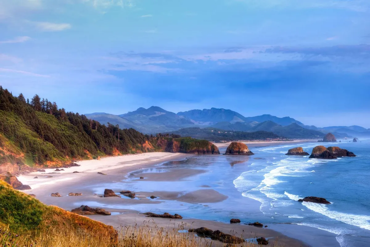 A wide, serene beach scene with ocean waves, striking rock formations, and a distant tree-lined backdrop. The natural elements evoke a sense of exploration and outdoor adventure, while the sandy stretch is ideal for family play, kite flying, and picnics.