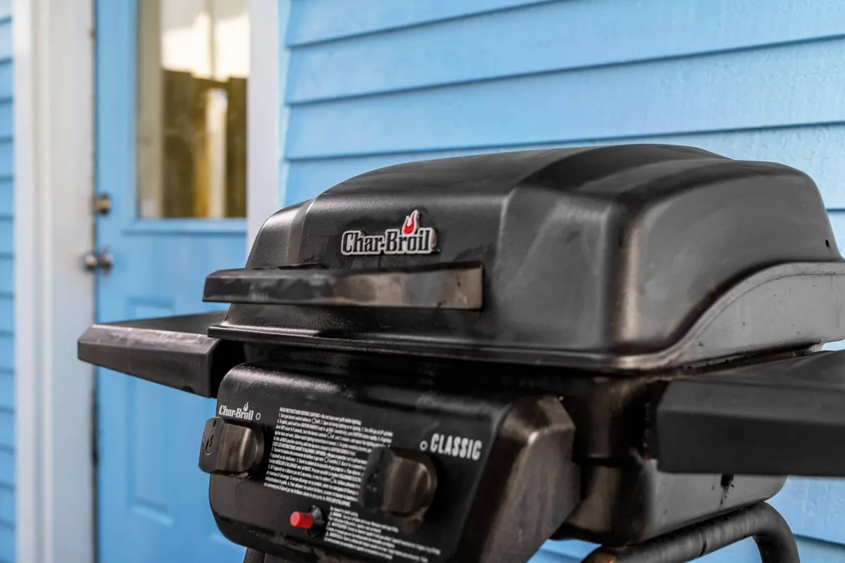 An exterior scene featuring a BBQ grill prominently placed on a patio or deck area with a blue door in the background. The space looks casual and inviting, ideal for family barbecues or gatherings with friends. The design signals a seamless indoor-outdoor connection.