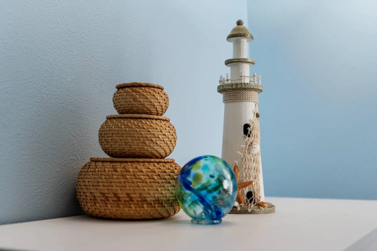 A serene coastal-themed living space featuring woven baskets, a lighthouse figurine, and a glass orb. The décor and accessories evoke feelings of the nearby beach and shoreline, tying the indoors to the coastal location.