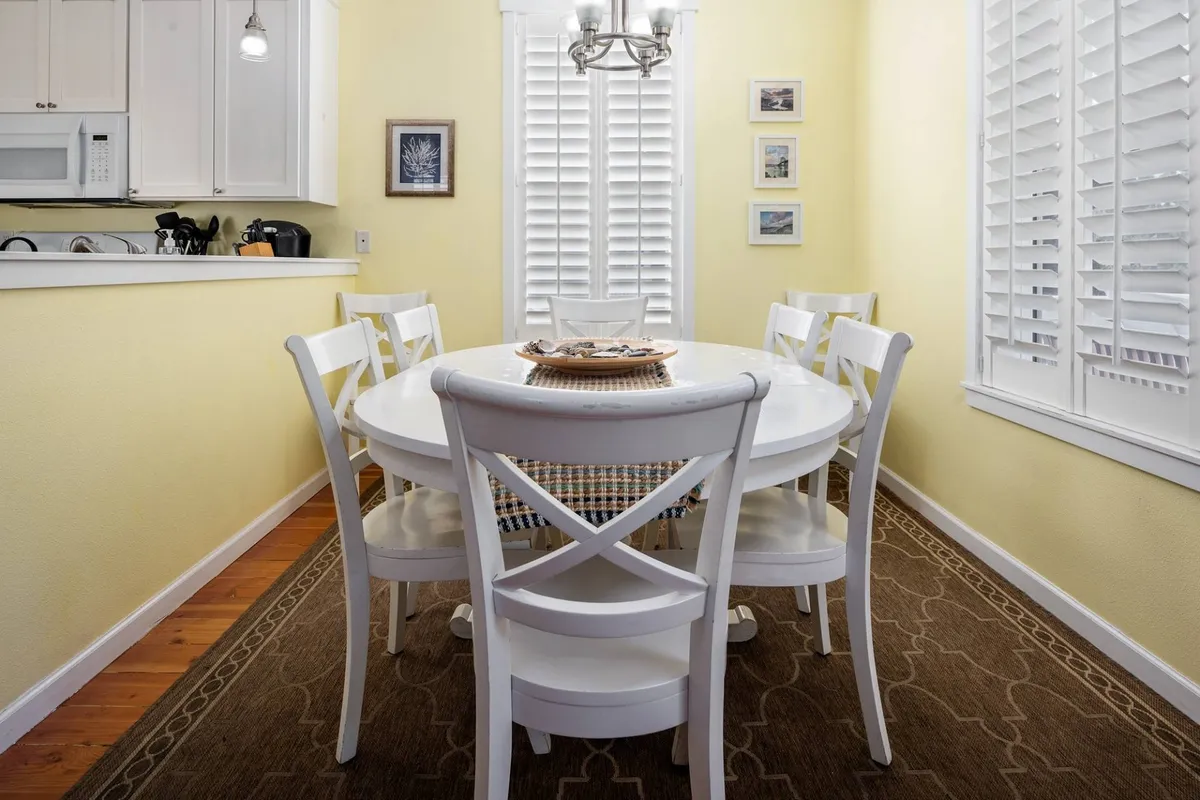 A charming dining area with a round farmhouse table, white chairs, a striking chandelier overhead, and natural light filtering through a shuttered window. The table is set for intimate family meals or festive gatherings.
