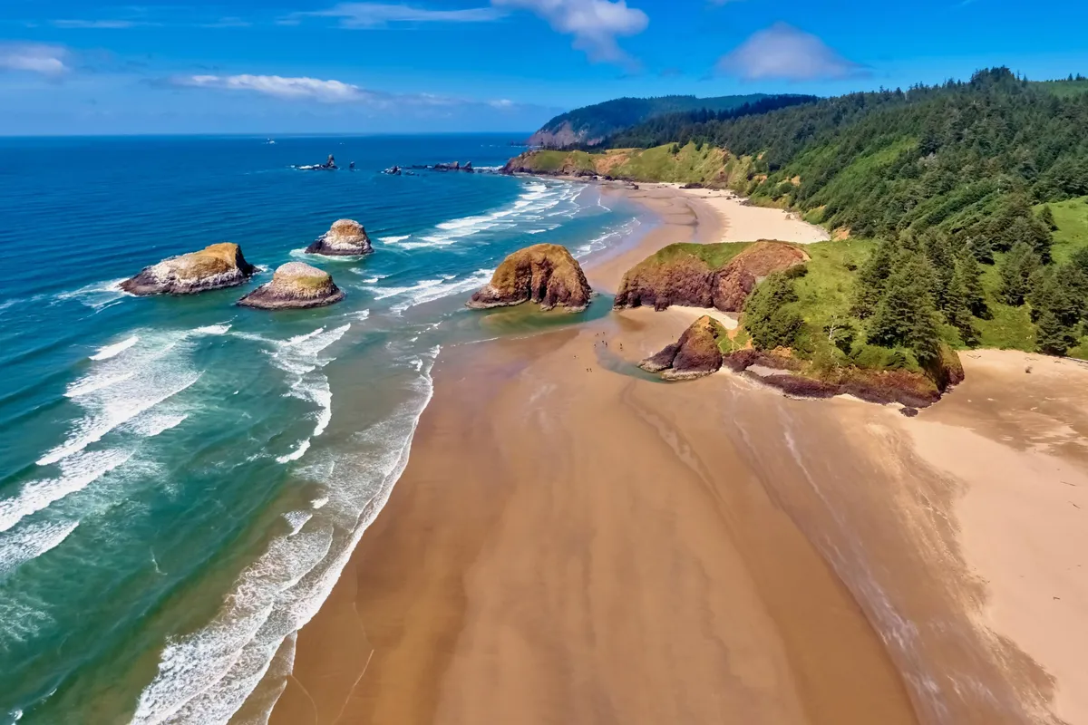 A tranquil beach view showing waves breaking on the sand, with unique rock formations and green hills in the distance. The scene suggests peaceful exploration, beachcombing, and the joy of discovering the natural beauty just steps from the neighborhood.
