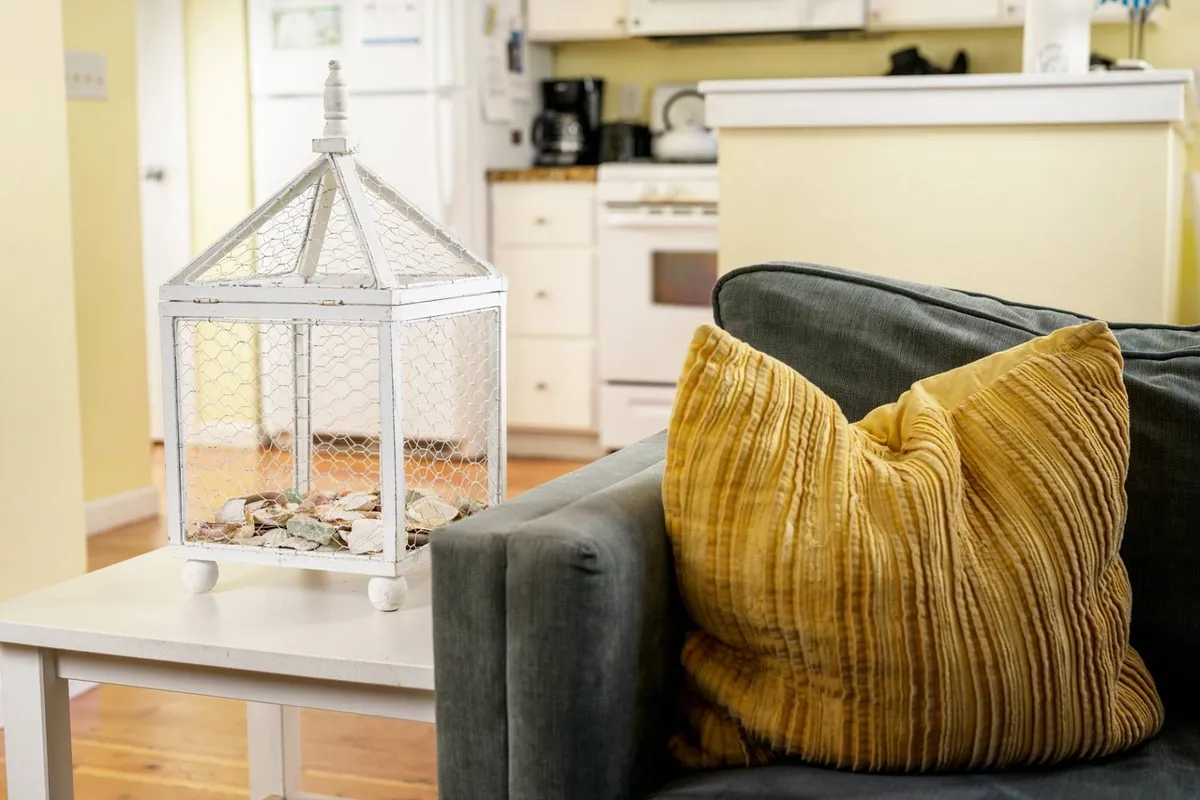A comfortable living area with a gray sofa, yellow textured pillow, and a white decorative lantern that creates a warm, inviting atmosphere. The setting suggests a cozy space for couples to relax, unwind, and plan their day of coastal exploration.