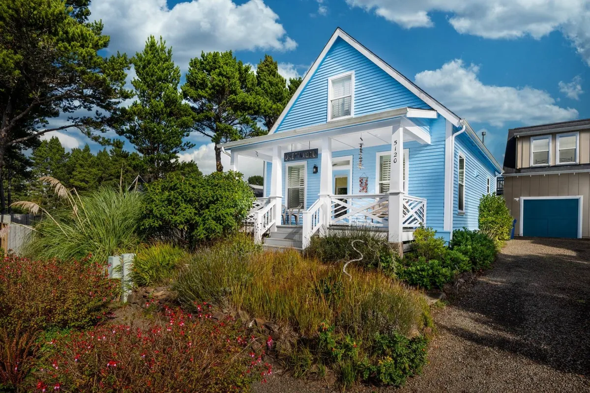The image showcases a charming blue cottage nestled in a tranquil neighborhood, with a welcoming front porch surrounded by a lush, colorful garden. The inviting porch hints at cozy mornings with coffee or relaxing sunset evenings, while the meticulously maintained landscaping enhances the sense of a private retreat.